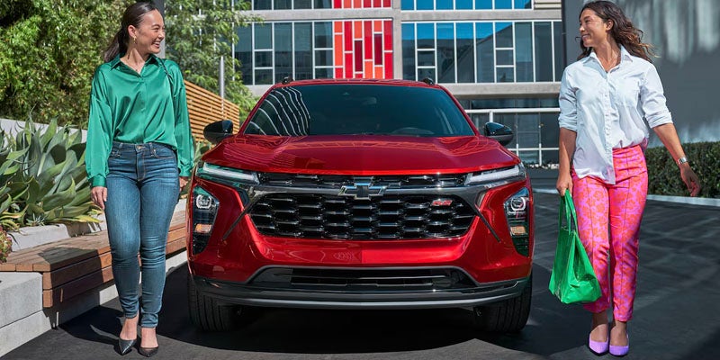Two women stand beside a red 2025 Chevrolet Trax, smiling and posing for the camera in a sunny outdoor setting.