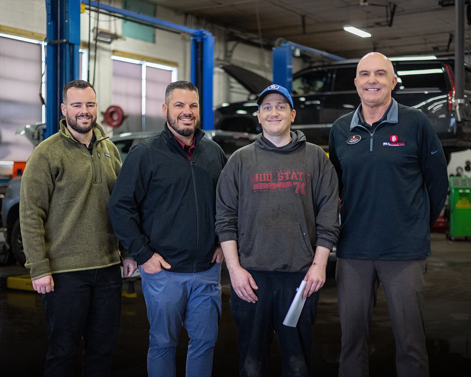 Three men wearing green shirts and blue jackets stand together, smiling in a casual outdoor setting.