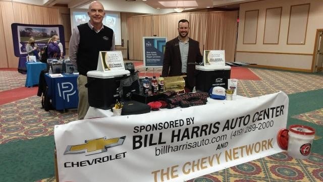 Two men stand behind a table displaying a Chevrolet logo, smiling and engaging with an audience.
