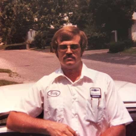 A man wearing a white shirt and glasses stands beside a parked car, smiling at the camera.