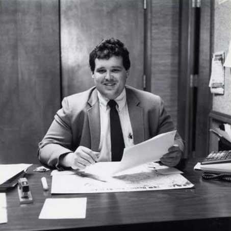A man in a suit and tie sitting at a desk, focused on his work with a laptop and documents in front of him.