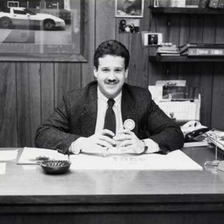 A man in a suit sits at a desk with a nameplate, focused on his work in a professional office setting.